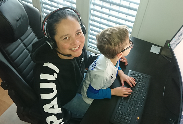 Windows Server Administratorin Sallo sitzt im Homeoffice mit Headset an ihrem Schreibtisch. Auf ihrem Schoß sitzt ihr Sohn und schaut auf den Bildschirm.